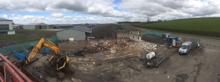 Digger carrying out a site clearance in front of a hangar with a pile of rubble behind the digger on the ground